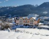 Hotel s bílou fasádou a balkony stojí na zasněženém svahu s horskou krajinou v pozadí.
