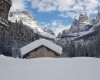 Horské chaty pokryté sněhem v zasněženém údolí s lesy a horami pod modrou oblohou.