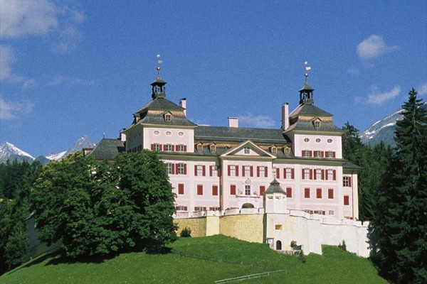 Historický zámek stojící na kopci obklopený stromy s horami v pozadí.