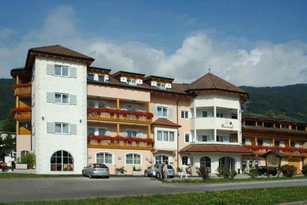 Hotel s balkony a květinami na přední straně, obklopený horami.