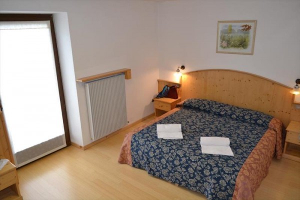 Ložnice s dvoulůžkem, nočními stolky, radiátorem a výhledem na balkonové dveře.
