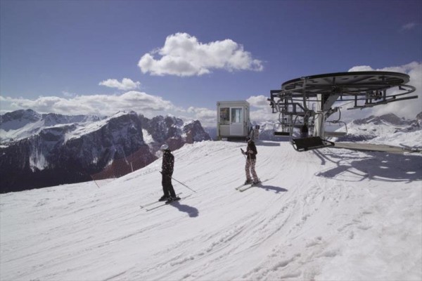 Dva lyžaři na zasněženém vrcholu vedle lanovky s horským panoramatem v pozadí.