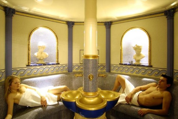 Dva lidé relaxují v luxusní parní lázni s mozaikovými zdmi a dekorativními prvky.  