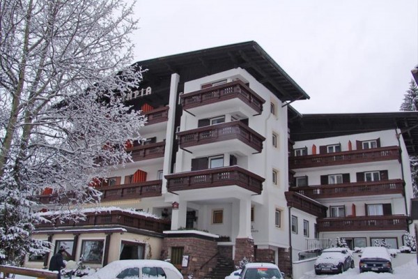 Čtyřpatrový hotel se sněhem pokrytými balkony a auty zaparkovanými před ním.
