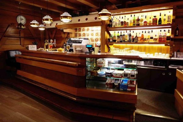 Dřevěný bar s osvětlenými policemi plnými lahví alkoholu a vitrínou se sladkostmi.