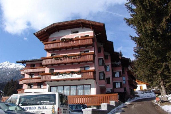 Horský hotel s dřevěnými balkony a bílým dodávkovým autem v popředí, zasněžené hory v pozadí.