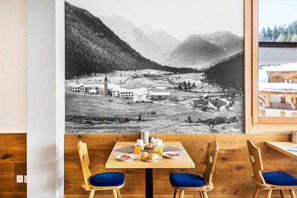 Stůl se snídaní v restauraci s velkou černobílou fotografií alpské krajiny na stěně.