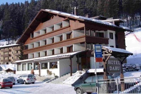 Dvoupatrový horský hotel na zasněžené krajině s parkovištěm a okolními stromy.