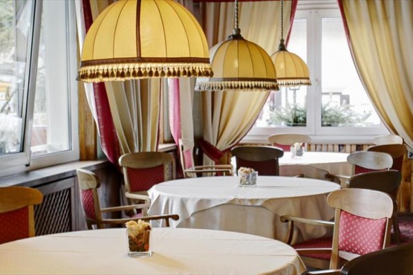 Útulná restaurace s kulatými stoly, červenými židlemi a závěsy, nasvětlená velkými lampami.