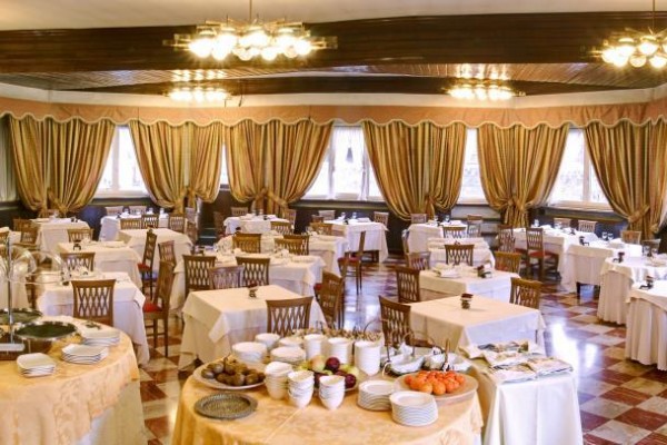 Útulná restaurace s kulatými stoly, červenými židlemi a závěsy, nasvětlená velkými lampami.