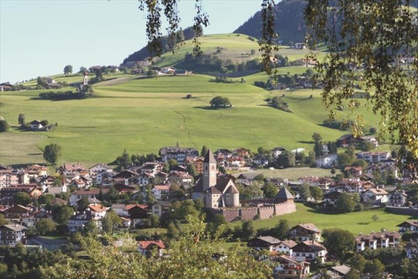 Středověký hrad obklopený domy v malebné krajině s kopci v pozadí.