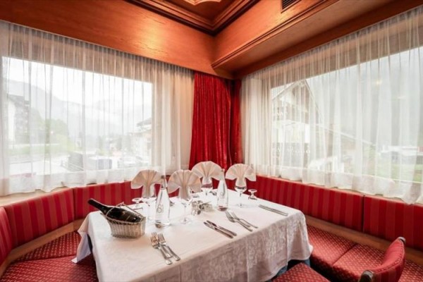Stůl pro šest osob s bílým ubrusem, slavnostně připravený v rohové restauraci s červeným závěsem a velkými okny.