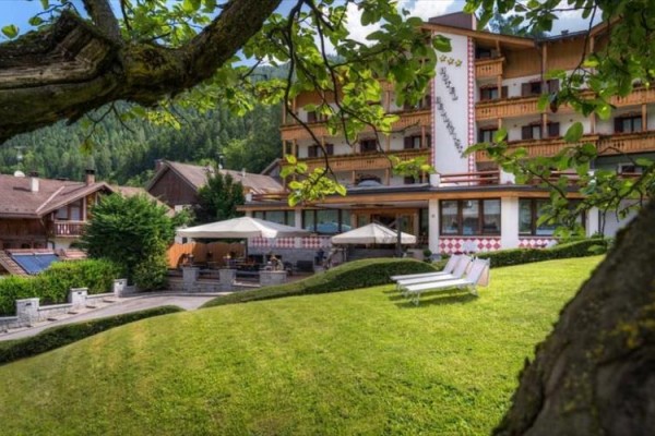 Hotel obklopený zelení s lehátky na trávníku a stromem v popředí.