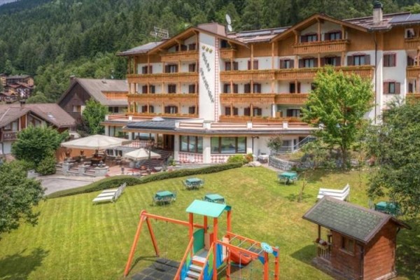 Hotel s terasou a dětským hřištěm na trávníku obklopený lesem.