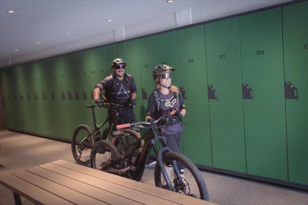 Dva cyklisté s přilbami tlačí kola kolem řady zelených skříněk.