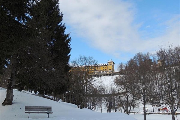 Sněhem pokrytý park s lavičkou a v dálce žlutá budova na kopci.