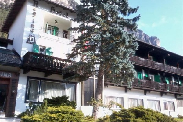 Hotel s balkony a zelenými okenicemi obklopený horami a stromy.