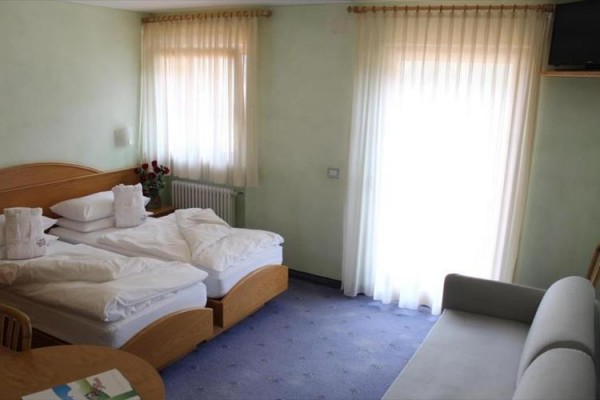 Světlý hotelový pokoj s dvěma postelemi, pohovkou a oknem s bílými závěsy.