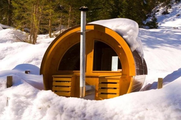 Dřevěná sudová sauna obklopená sněhem v lesním prostředí.