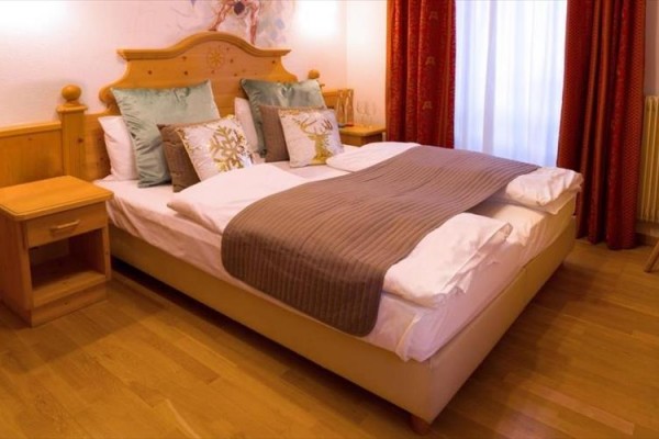 Dvoulůžková postel s polštáři a přehozem na dřevěné podlaze v pokoji s červenými závěsy.
