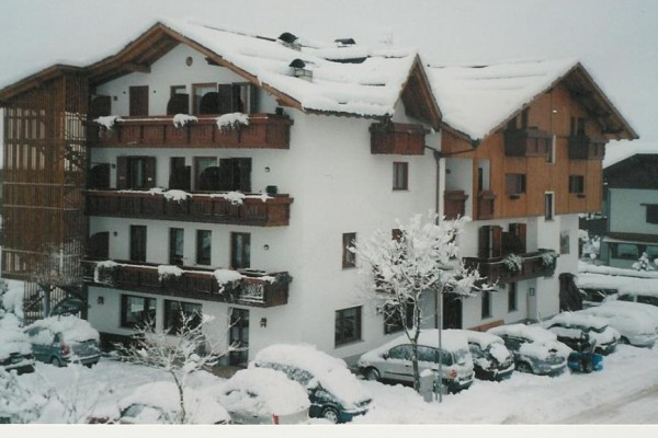 Zasněžený alpský hotel s balkony a zaparkovanými auty.