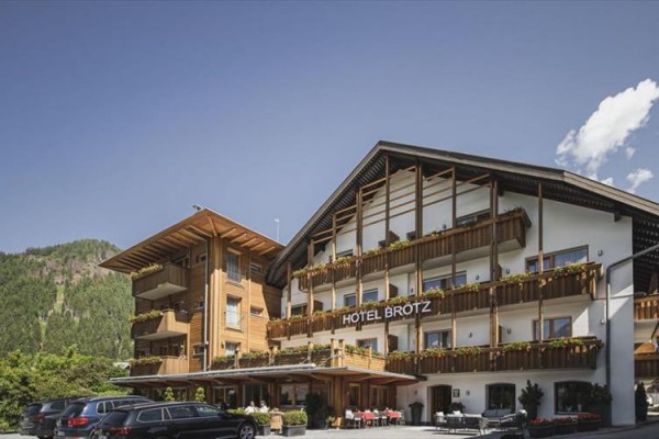 Budova hotelu s dřevěnými balkony a zaparkovanými auty před ním, v pozadí les a modrá obloha s mraky.