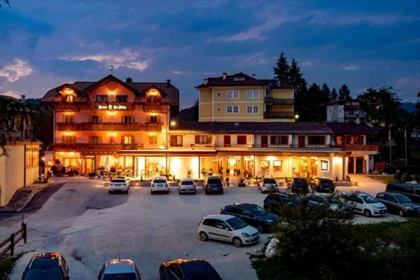 Hotel s osvětlenou fasádou a parkovištěm plným aut za soumraku.  