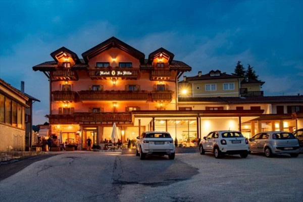 Večerní pohled na osvětlený hotel s parkovištěm a auty před ním.