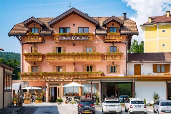 Třípatrová růžová budova hotelu s dřevěnými balkony a zaparkovanými auty před ní.  