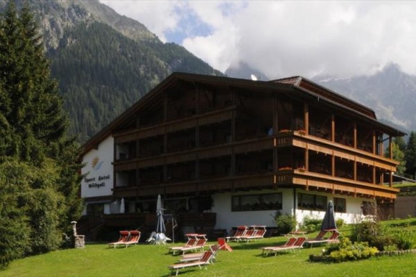 Dřevěný hotel v alpské krajině s lehátky na travnaté ploše.