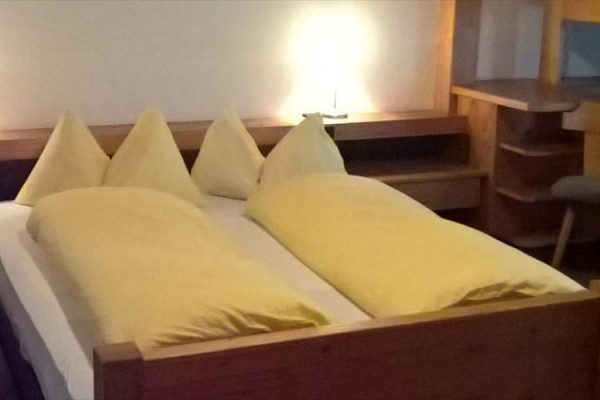 Manželská postel s dřevěným rámem a žlutým povlečením, vedle nočního stolku se svítící lampičkou.  