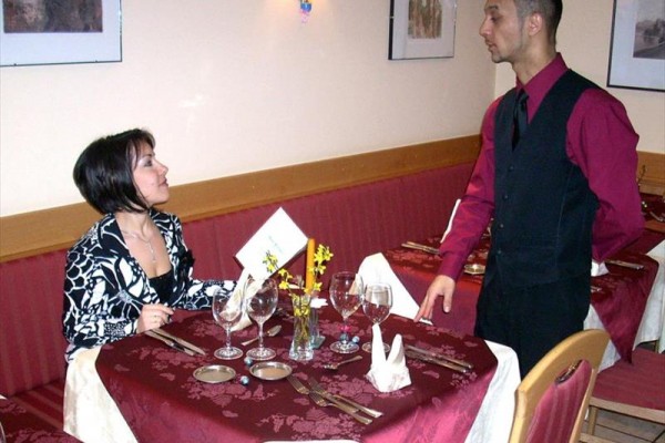 Žena sedící u stolu v restauraci diskutuje s číšníkem, který stojí vedle.