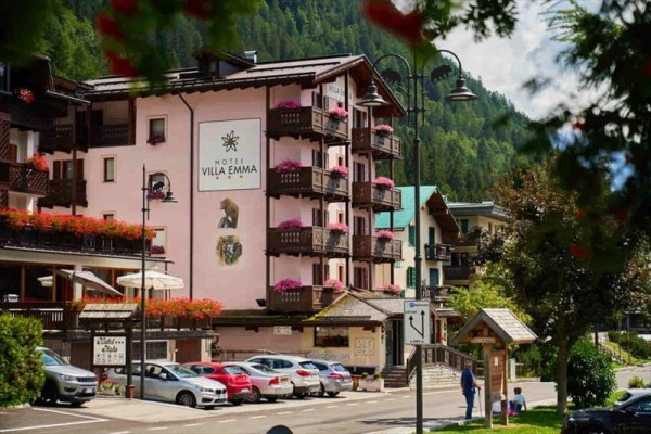 Růžový hotel Villa Emma s květinami na balkonech vedle zaparkovaných aut v horské oblasti.