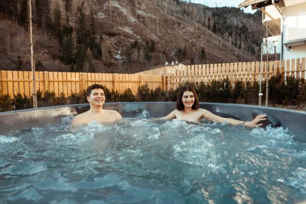 Dva lidé relaxují ve venkovní vířivce s dřevěným plotem a horami v pozadí.
