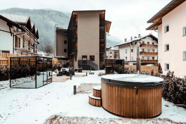Dvůr hotelu s venkovní vířivkou pokrytý sněhem obklopený budovami v horské krajině.