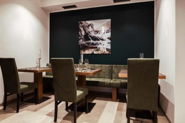 Restaurace s dřevěnými stoly a zelenými židlemi proti tmavé zdi s obrazem hor.