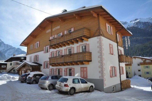 Horské ubytování s balkóny, zasněžené parkoviště a auta, v pozadí hory.