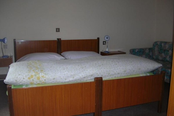 Dřevěná postel se dvěma lampičkami na nočních stolcích a květovanou pohovkou v pozadí.