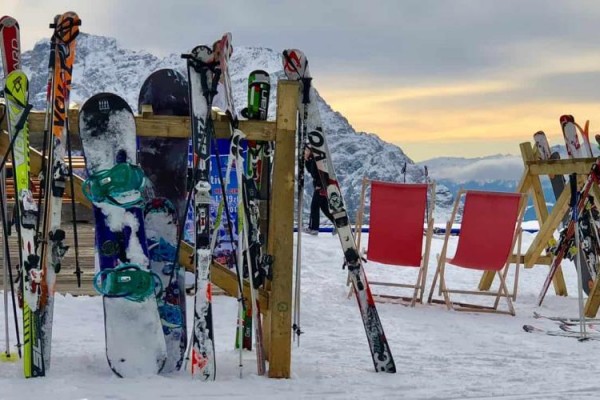 Lyže a snowboardy opřené o stojany na zasněžené sjezdovce s horami v pozadí.