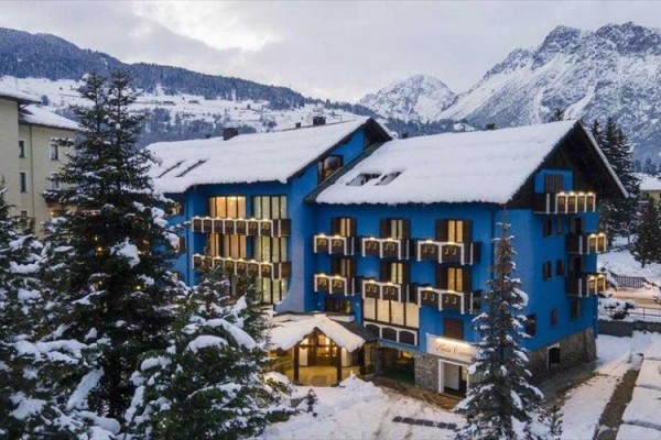 Modrý hotel obklopený zasněženými horami a stromy.