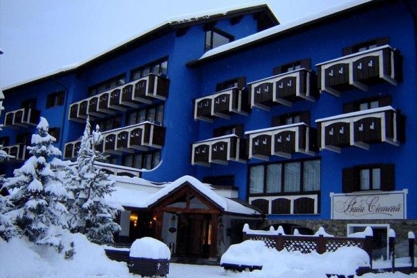 Modrá budova hotelu obklopena květinami na balkonech a terasou s lehátky a stolem.