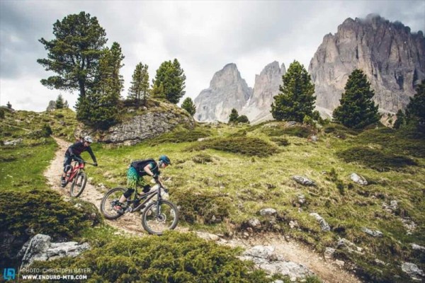 Dva cyklisté sjíždějí horskou stezku obklopenou stromy a skalami.