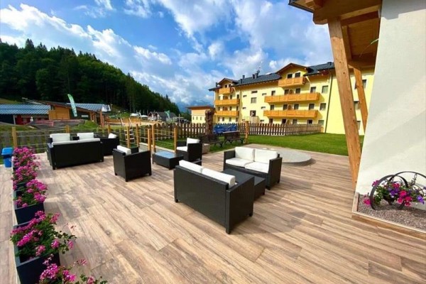 Terasa s ratanovým nábytkem, květinami a v pozadí žlutý hotel u lesa.