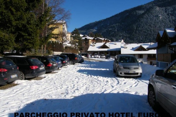 Sněhem pokryté parkoviště s auty u hotelu obklopeného horami.  