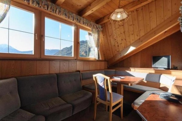 Dřevěný podkrovní obývací prostor se sedací soupravou, jídelním stolem a televizí, s výhledem na hory.