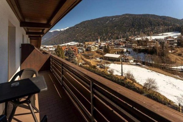Pohled z balkonu na zasněženou vesnici a okolní hory.
