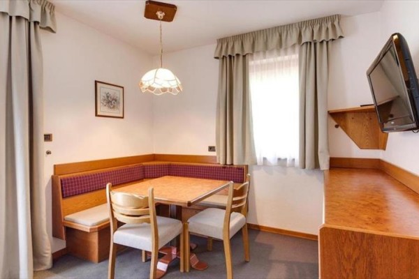 Jídelní kout s rohovou lavicí, dřevěným stolem a třemi židlemi v místnosti s televizí na zdi.