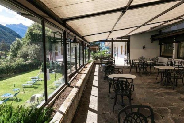 Zastřešená terasa s kovovými stoly a židlemi vedle zahrady s lehátky.