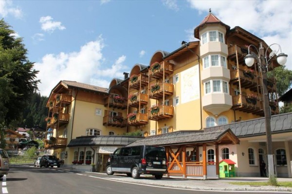 Žlutá budova ve stylu alpského hotelu s dřevěnými balkony a několika auty zaparkovanými před ní.