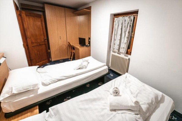 Dva jednolůžkové postele s ručníky, dřevěná skříň a pracovní stůl s počítačem v pokoji.
